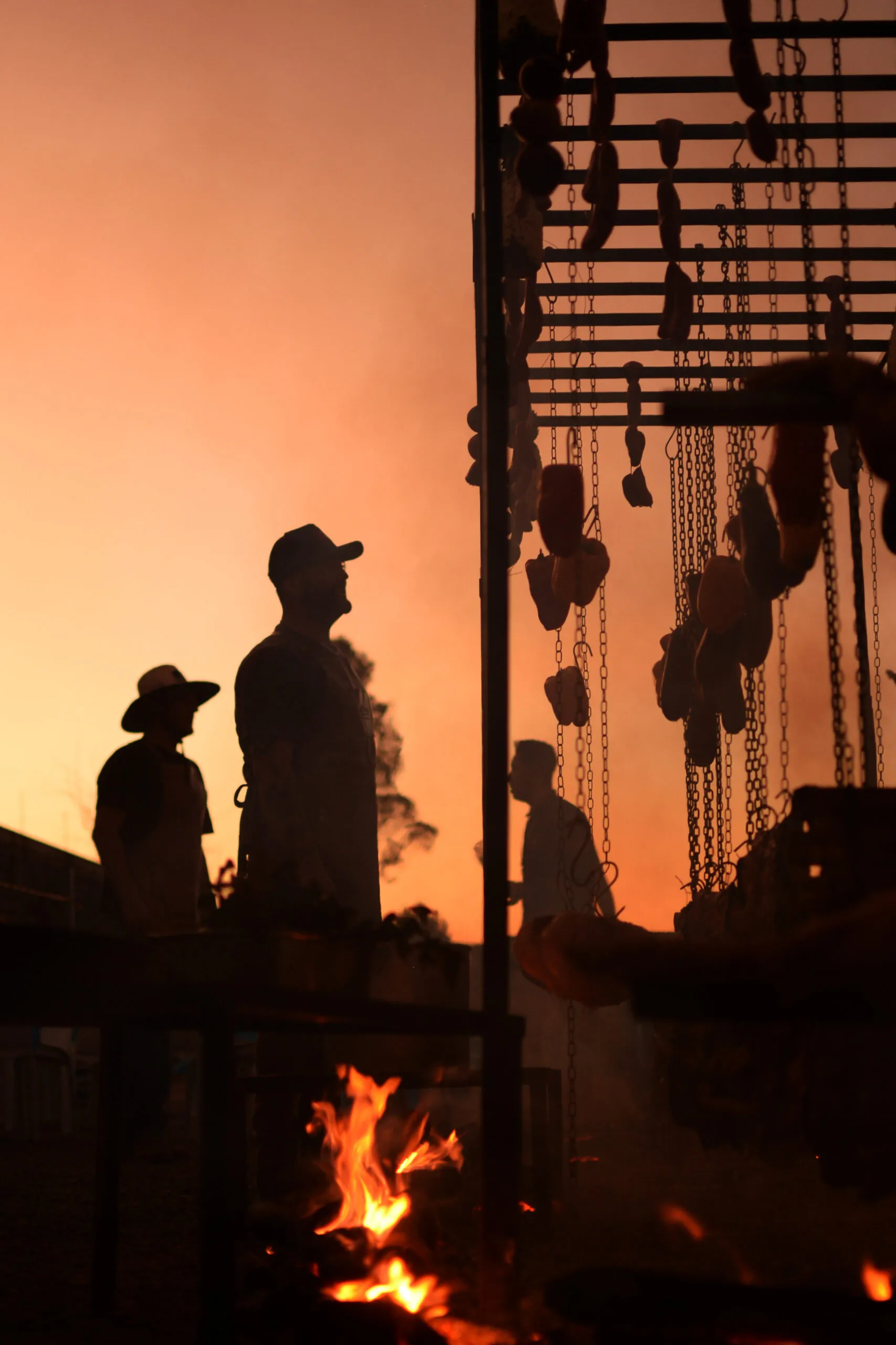 Silhuetas junto à estrutura do churrasco, com fogo e o pôr do sol ao fundo